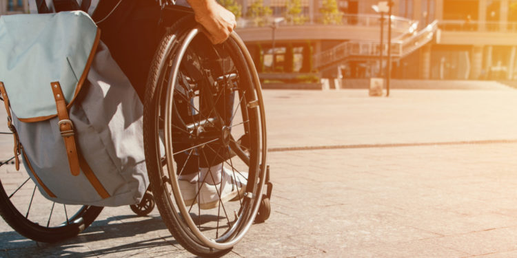 Cropped panoramic view of man using wheelchair with bag on street with sunlight